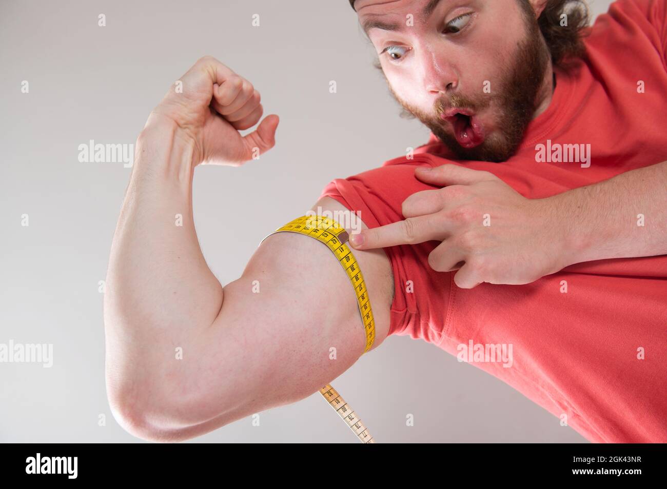 Bearded uomo divertente che misura bicipiti, muscoli del suo braccio con un metro a nastro giallo. Sorride, felice. Indossare una maglietta rossa. Sfondo grigio chiaro. Foto Stock