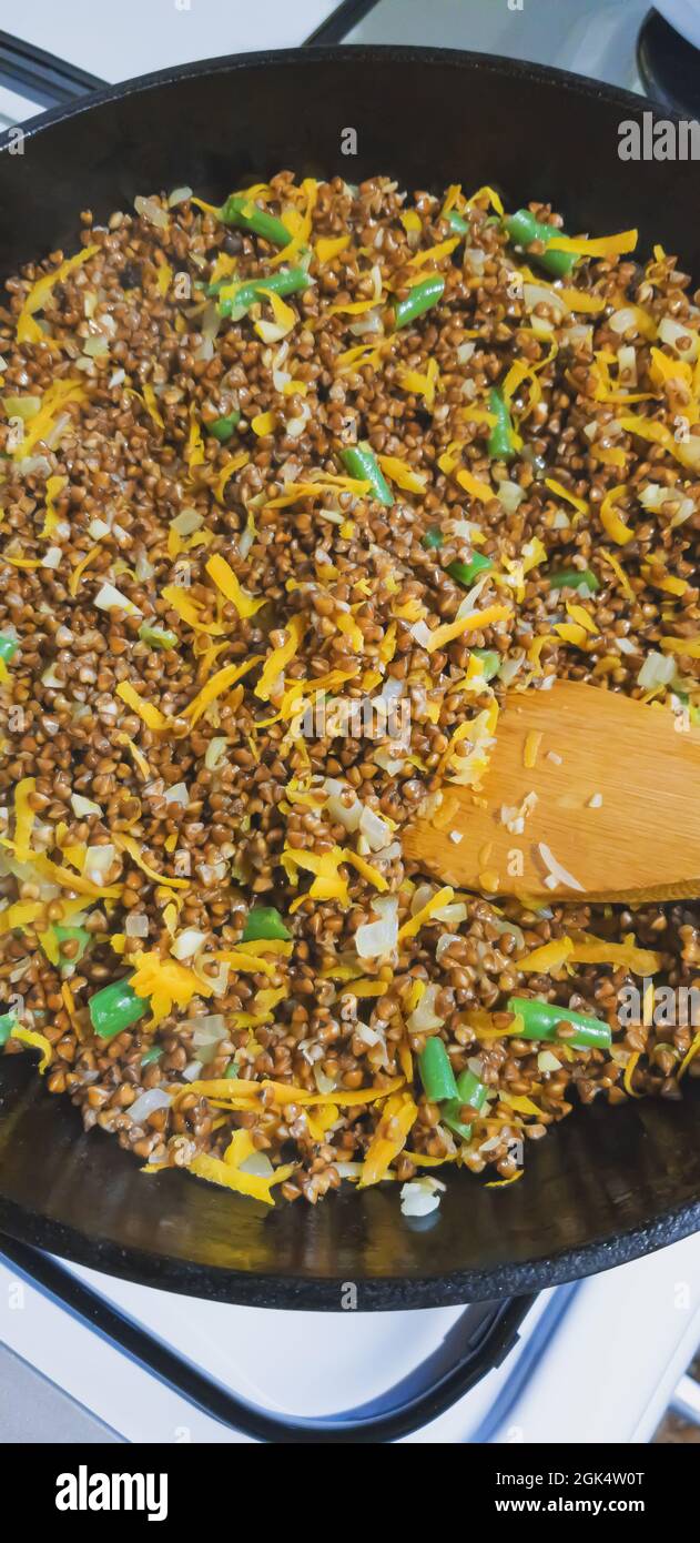 Cottura del grano saraceno con cipolle, carote, fagioli di asparagi in una padella di ghisa profonda su un forno a gas, con un cucchiaio di legno per mescolare. Foto Stock