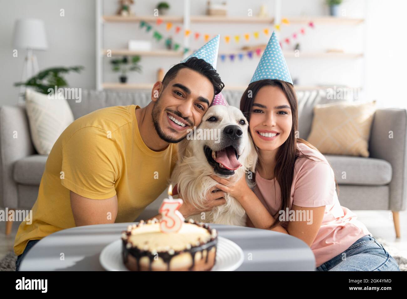 Felice coppia multirazziale celebrare il compleanno del loro cane con torta di festa, portando i cappelli di festa a casa Foto Stock