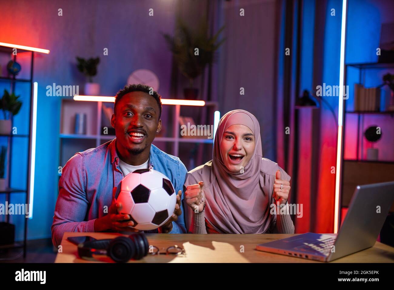 Ritratto di uomo africano e donna musulmana emotivamente allietare la squadra preferita durante la partita di calcio. Giovane coppia che usa un moderno notebook per guardare la partita a casa. Foto Stock