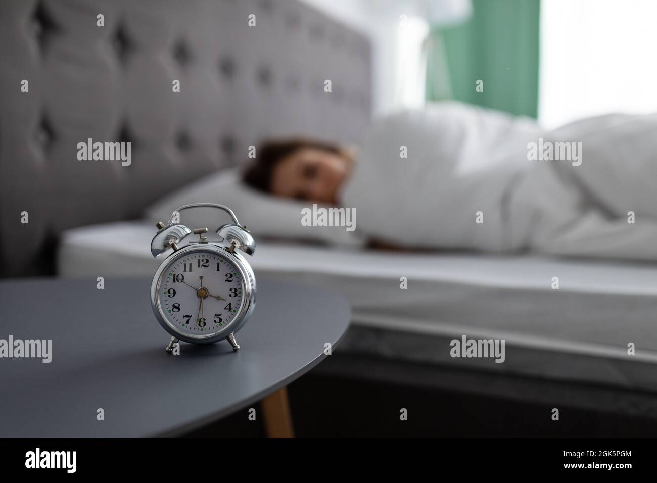 Sveglia sul comodino e vista offuscata della giovane donna sdraiata a letto, coperta di coperta, non volendo svegliarsi Foto Stock