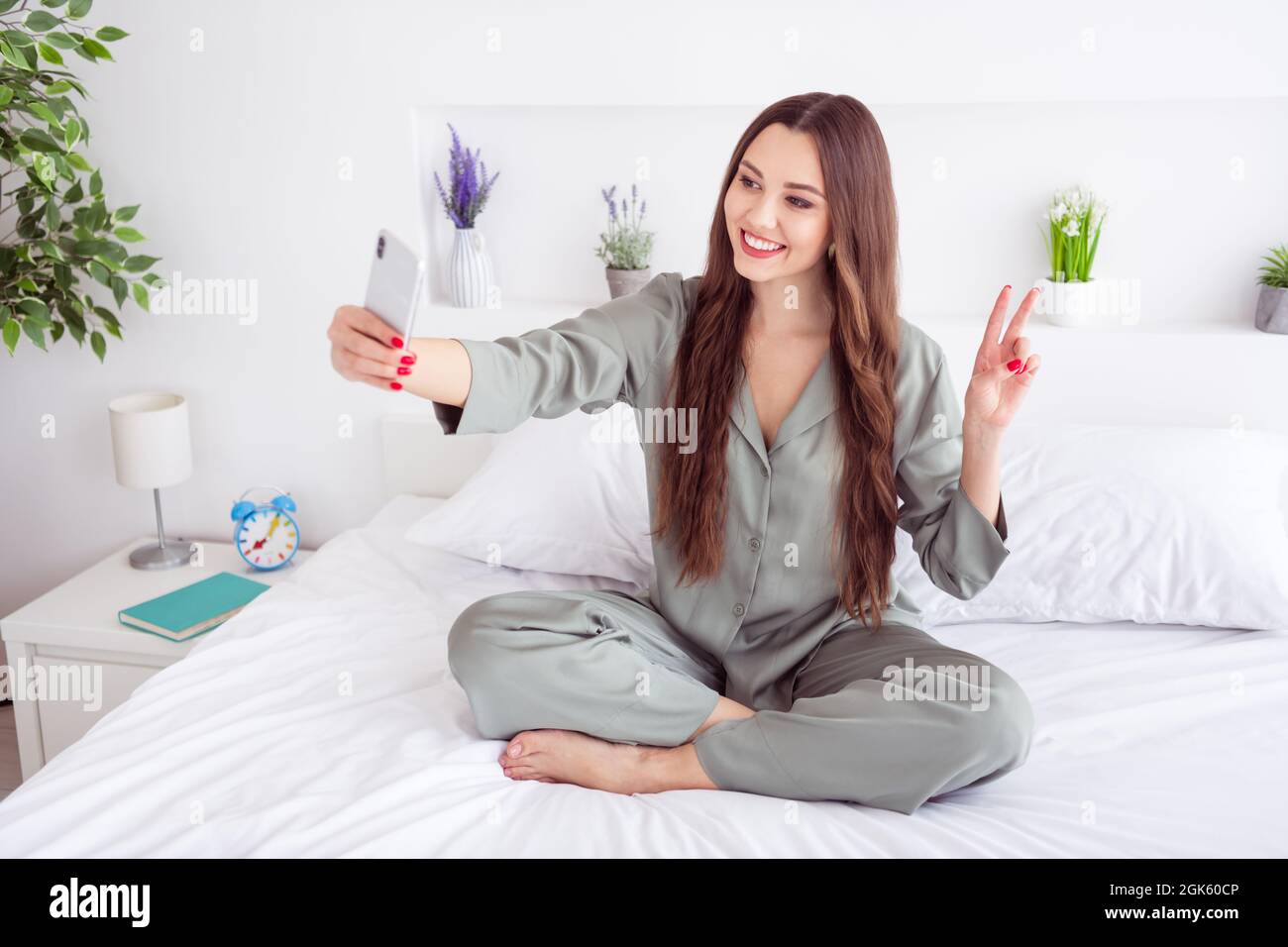 Foto di adorabile signora grigio sleepwear seduta letto gambe incrociate  prendendo selfie gadget moderno mostrando V-segno interno stanza casa Foto  stock - Alamy