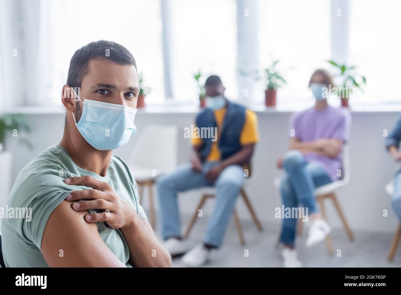 giovane uomo in maschera medica guardando la macchina fotografica vicino sfocato persone multietniche nel centro di vaccinazione Foto Stock