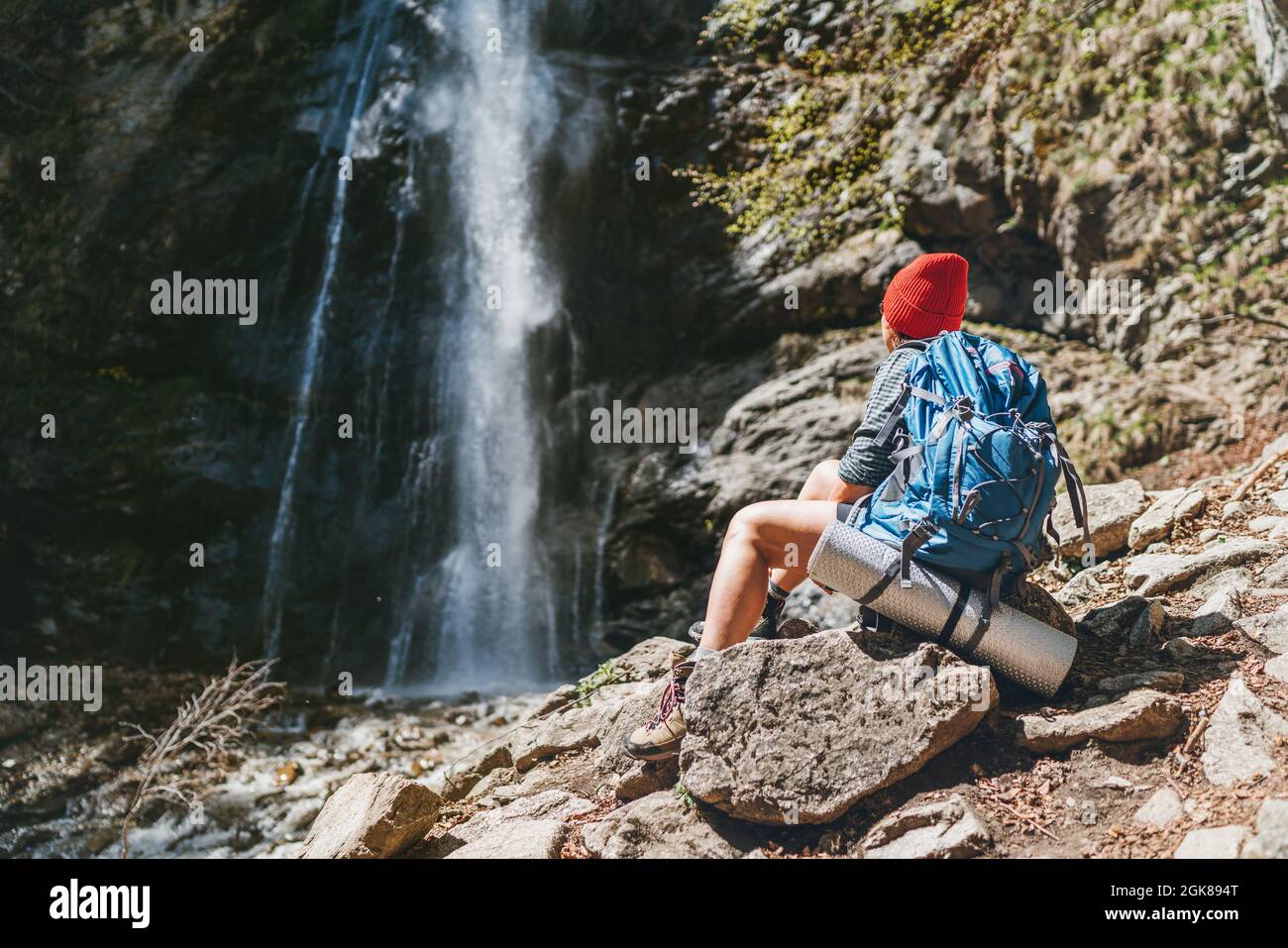 Donna con uno zaino in cappello rosso vestito con abiti da trekking attivi seduta vicino alla cascata del fiume montagna e godendo del potere della natura. Foto Stock