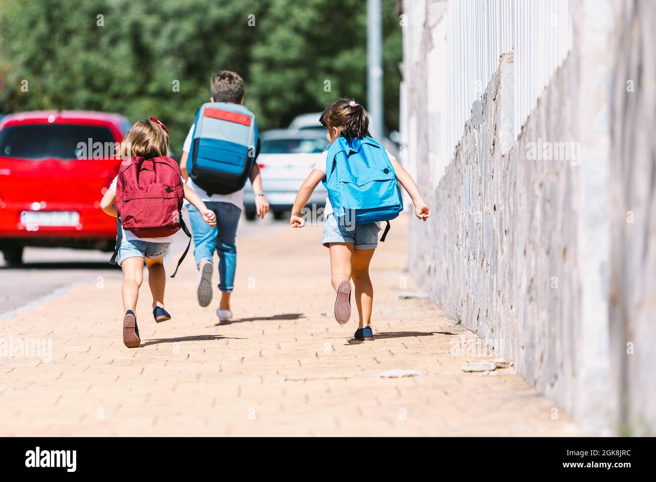 Vista posteriore di anonimi scolari con zaini che corrono su un vialetto piastrellato in una città soleggiata su sfondo sfocato Foto Stock