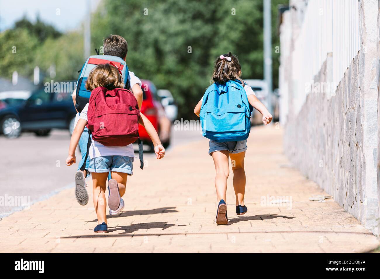 Vista posteriore di anonimi scolari con zaini che corrono su un vialetto piastrellato in una città soleggiata su sfondo sfocato Foto Stock