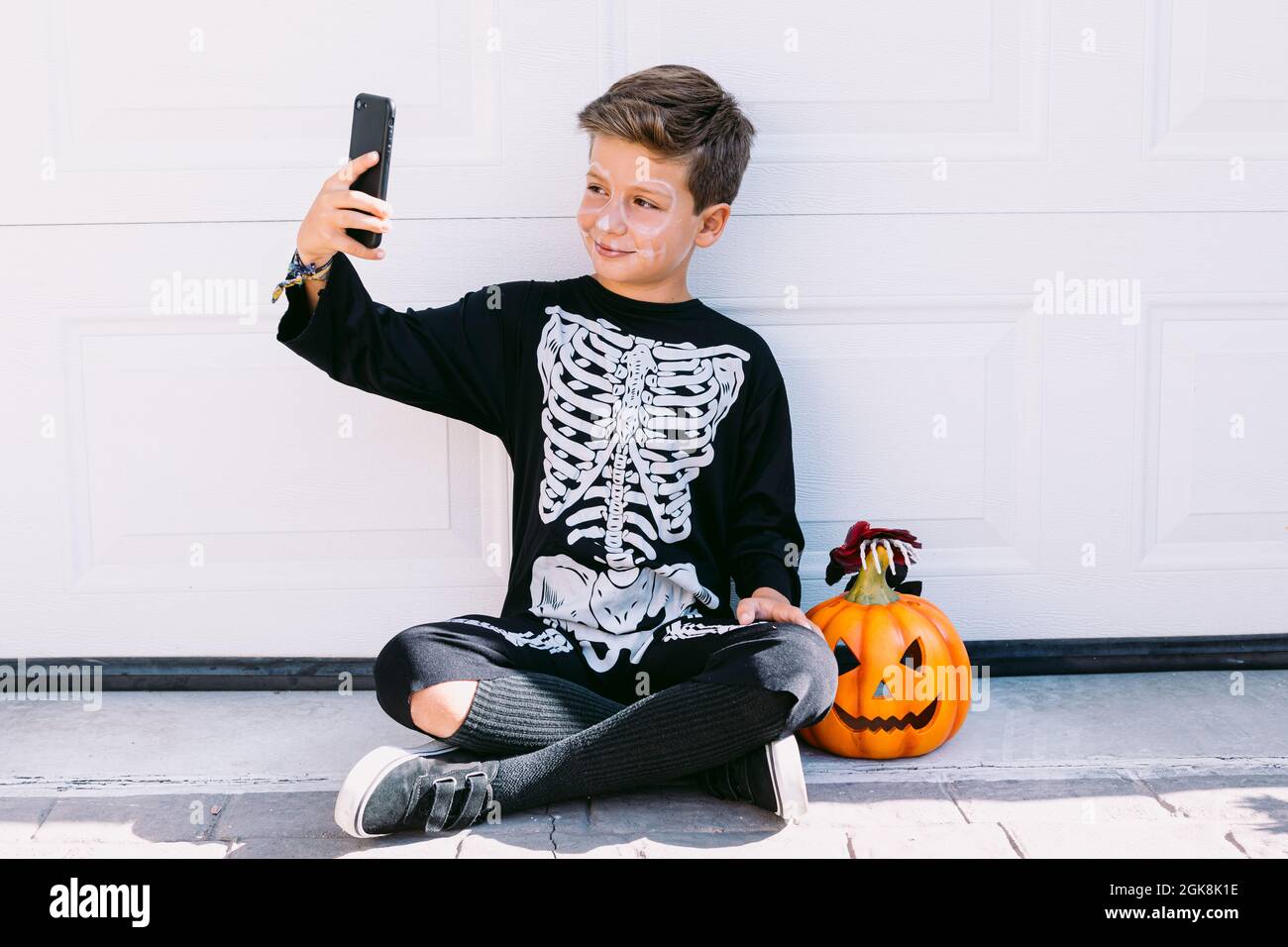 Ragazzo sorridente in costume di scheletro nero con faccia dipinta seduta vicino alla zucca di Halloween intagliata e prendendo selfie sullo smartphone vicino a parete bianca su tree Foto Stock