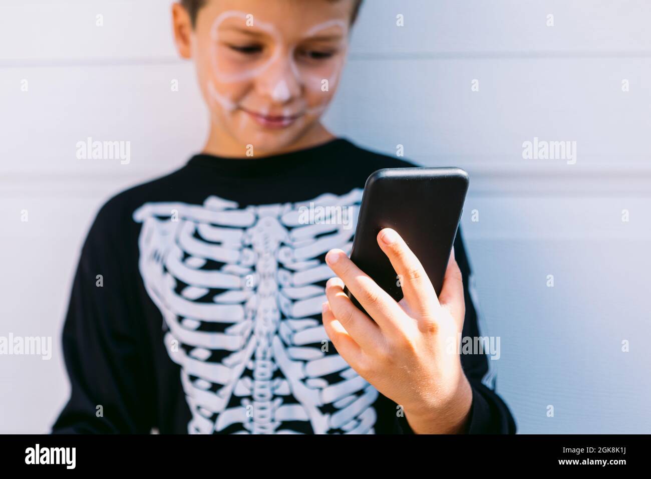Ragazzo allegro in costume di scheletro nero con volto dipinto utilizzando sul telefono cellulare mentre si siede vicino alla parete bianca sulla strada Foto Stock