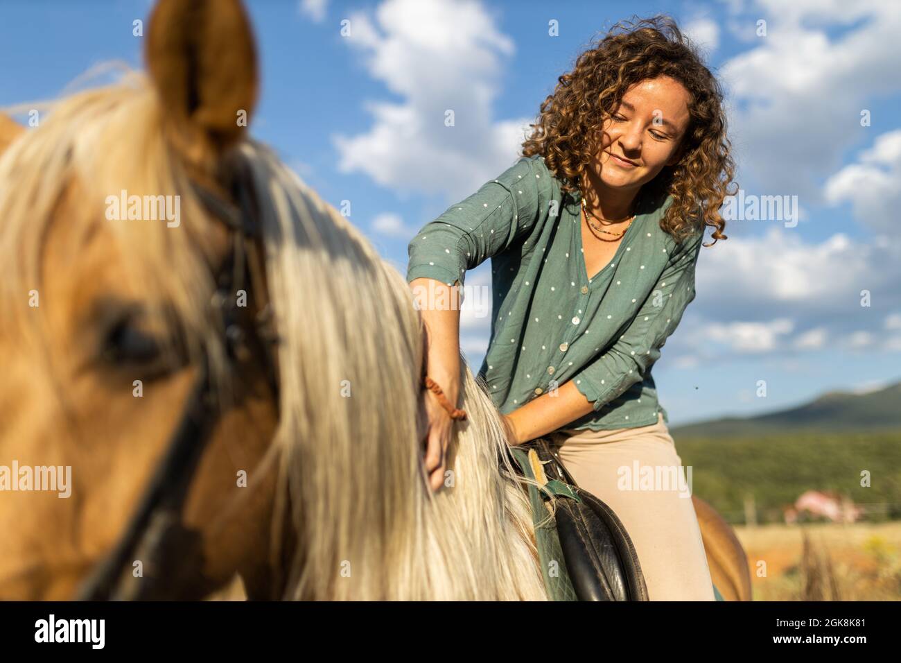 Donna sorridente con capelli ricci carezzando stallone mane sotto cielo nuvoloso blu alla luce del sole Foto Stock