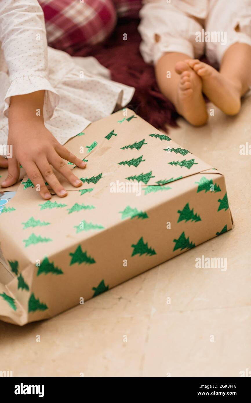 Crop anonymous bambino apertura presente scatola con abete modello sul pavimento durante Capodanno vacanza in casa Foto Stock