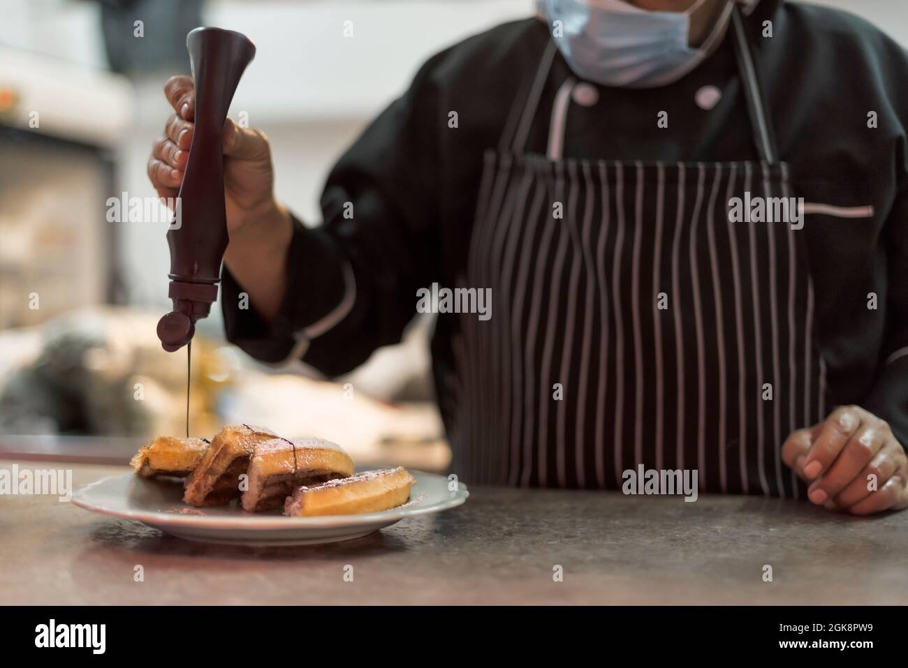 Raccolto irriconoscibile cuoco maschile in maschera sterile decorando deliziosi waffle viennesi con salsa di cioccolato da bottiglia in cucina ristorante Foto Stock