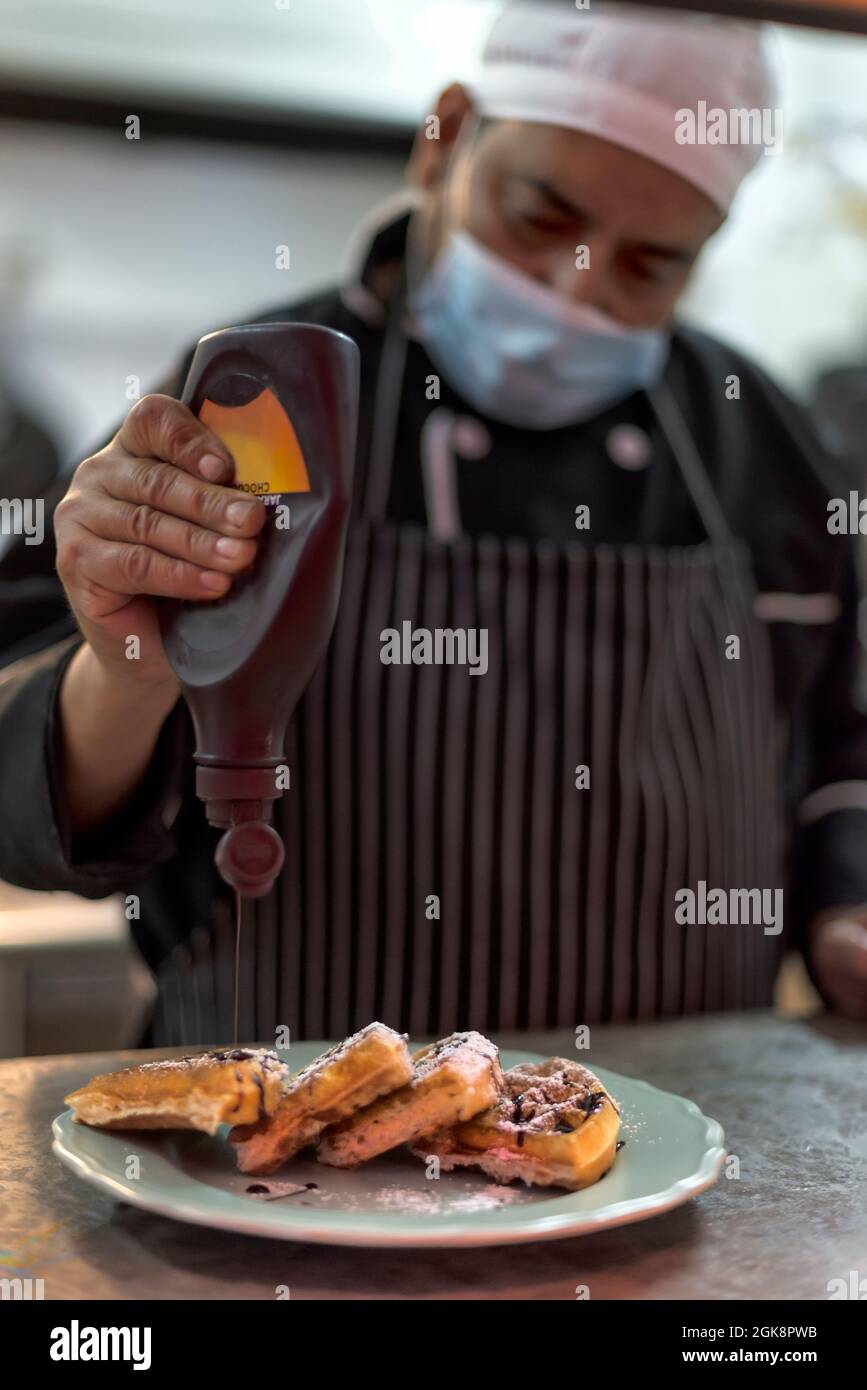 Raccolto etnico maschio cuoco in maschera sterile decorando deliziosi waffle viennesi con salsa di cioccolato da bottiglia in cucina ristorante Foto Stock
