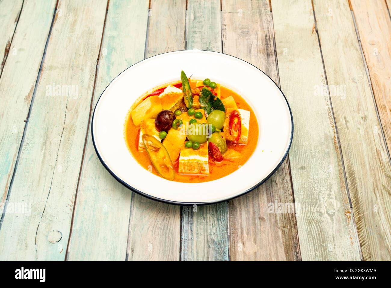 Curry rosso speziato thailandese con tofu e frutta su un piatto di metallo smaltato bianco e bordo blu Foto Stock