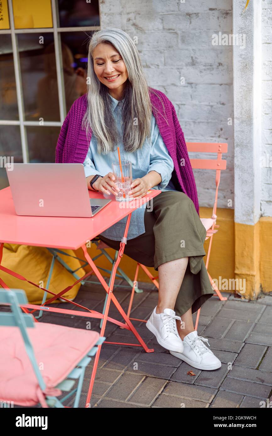 Sorridente donna asiatica con vetro d'acqua siede al tavolo di corallo con un laptop aperto Foto Stock