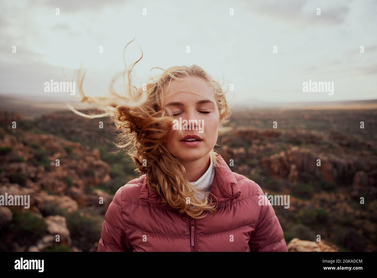 Giovane donna con occhi chiusi che sente vento fresco contro il viso in piedi sulla collina di montagna Foto Stock