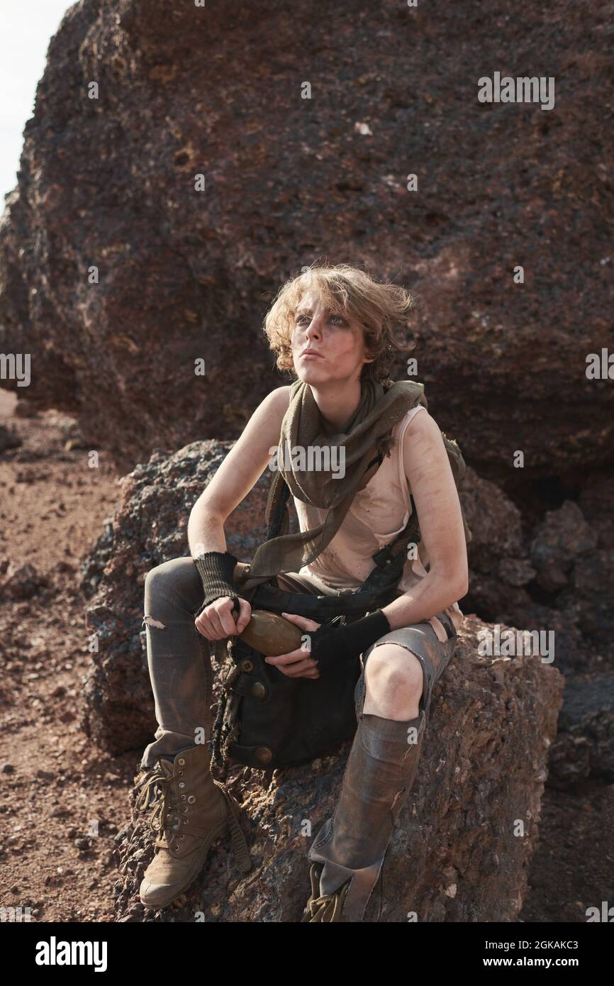 Giovane donna con il volto sporco che riposa sulla pietra nel deserto e beve acqua Foto Stock