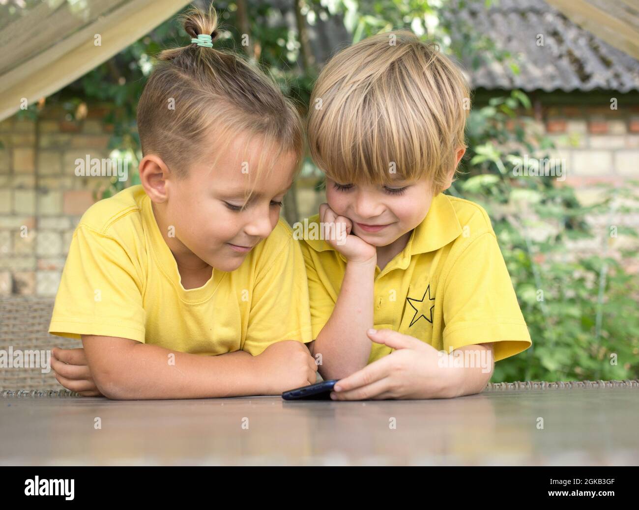 Carino 5 anni ragazzi in giallo T-shirt sono guardare lo schermo del telefono di fronte a loro con entusiasmo. Bambini moderni, era digitale, curiosità, passione Foto Stock