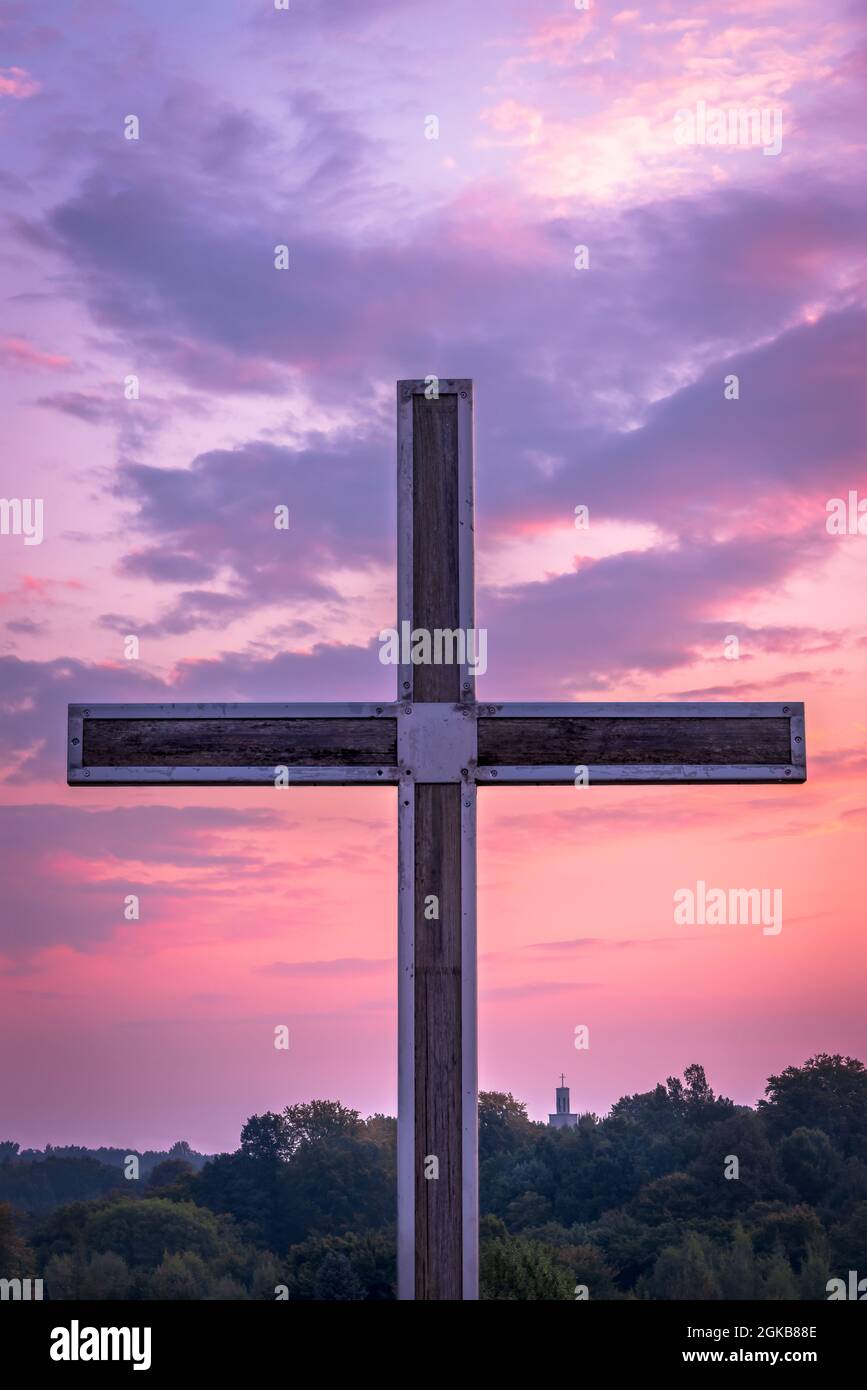 Croce cristiana in legno in un telaio di acciaio. Foresta e chiesa sullo sfondo. Luce del mattino, bello, colorato, cielo nuvoloso. Foto Stock