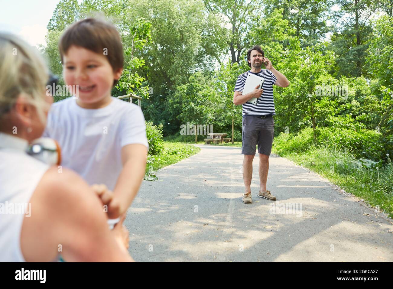 Madre e figlio in vacanza estiva nel parco con padre sul cellulare in background Foto Stock