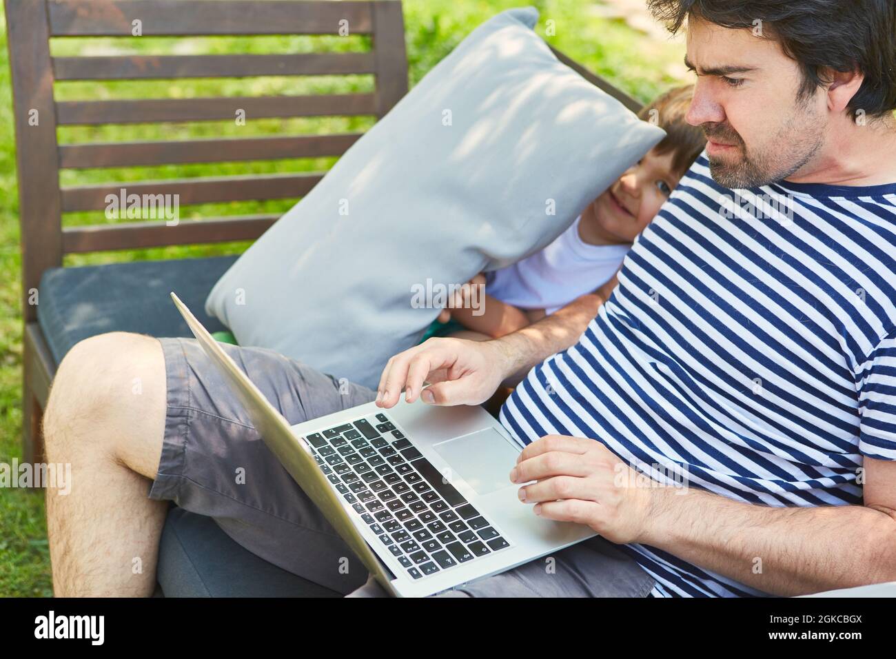 Il bambino disturba il padre sul PC portatile durante una videoconferenza in giardino durante le vacanze estive Foto Stock