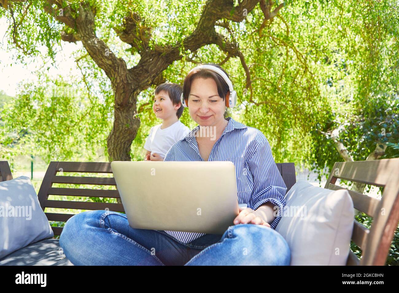 Donna su PC portatile durante una videoconferenza nel giardino verde con figlio in background Foto Stock