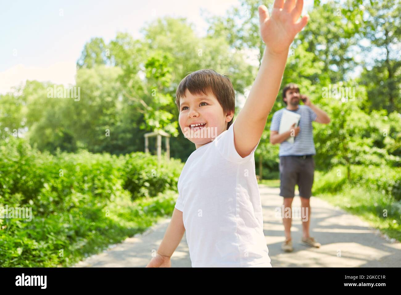 Padre che parla al telefono cellulare nel parco in estate con il figlio che gioca in primo piano Foto Stock