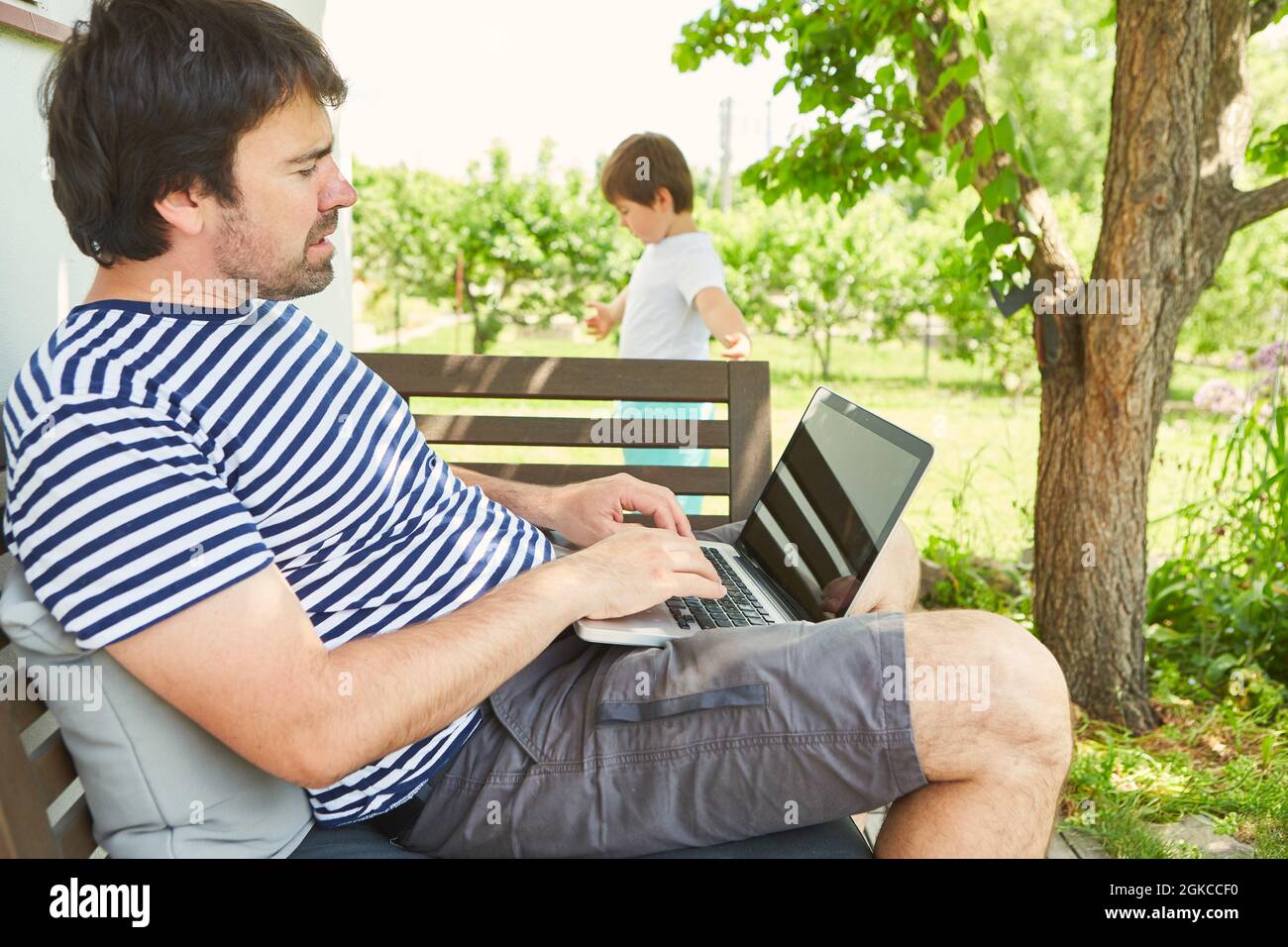 Uomo che lavora su un computer portatile in giardino in estate con bambino in background Foto Stock
