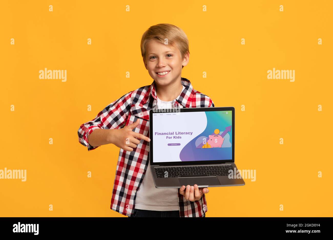 Ragazzo caucasico sorridente che punta allo schermo del laptop con il sito della scuola di alfabetizzazione finanziaria per bambini, sfondo arancione Foto Stock