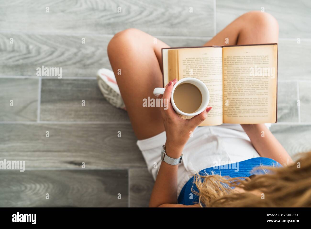 La giovane ragazza intellettuale si siede a terra e beve una tazza di caffè, mentre sta leggendo un libro. Foto di alta qualità Foto Stock