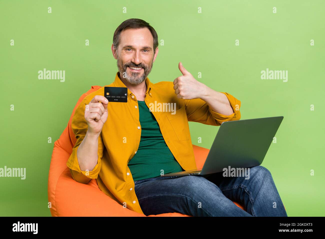 Foto di uomo matura bruna felice fare pollice su carta di credito laptop indossare casual vestito sedia seduta isolato su sfondo verde colore Foto Stock