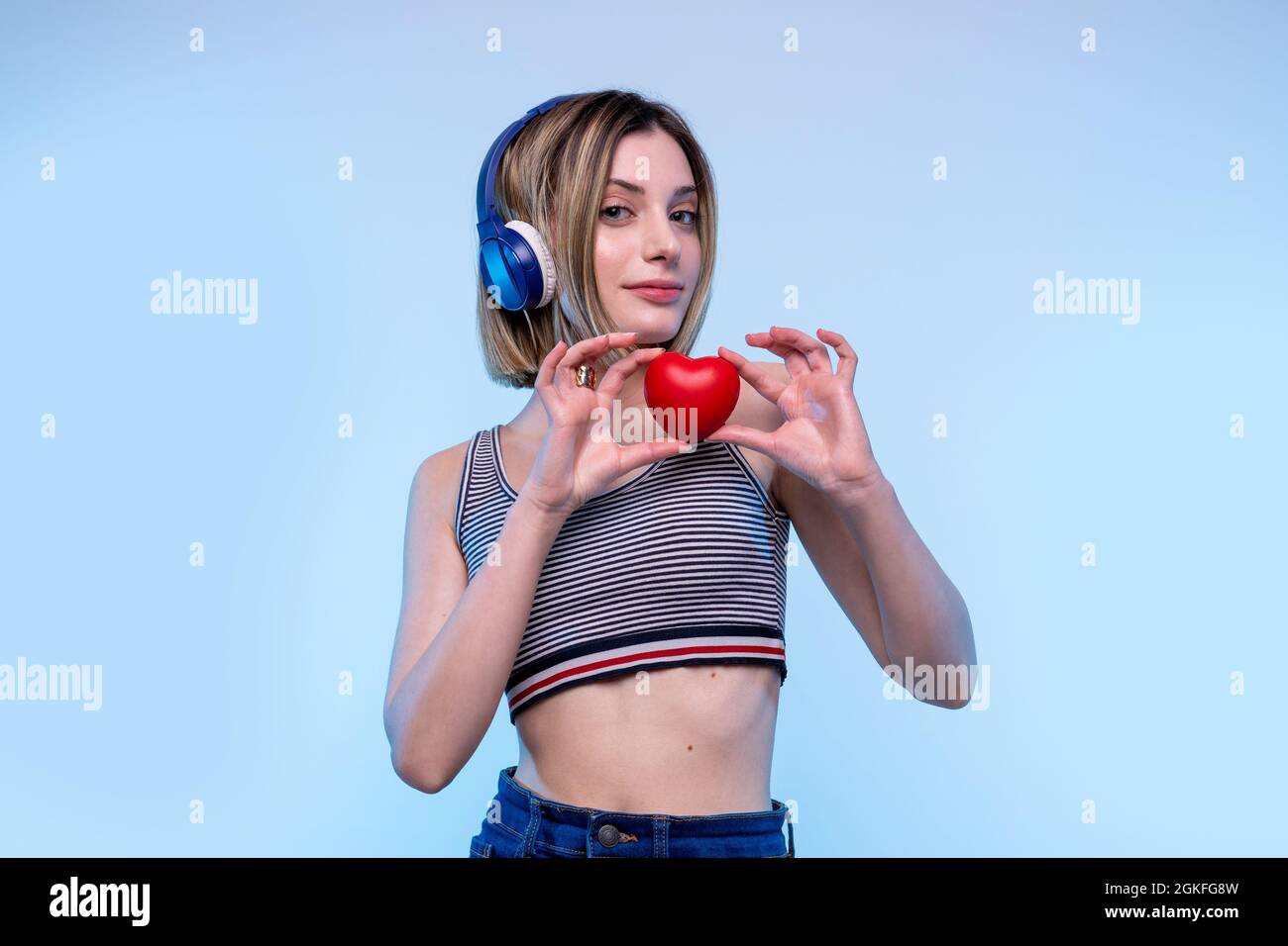 Giovane ragazza caucasica moderna che posa in piedi ascoltare musica con cuffie con uno sfondo blu chiaro - Studio fotografia di bella modella donna Foto Stock