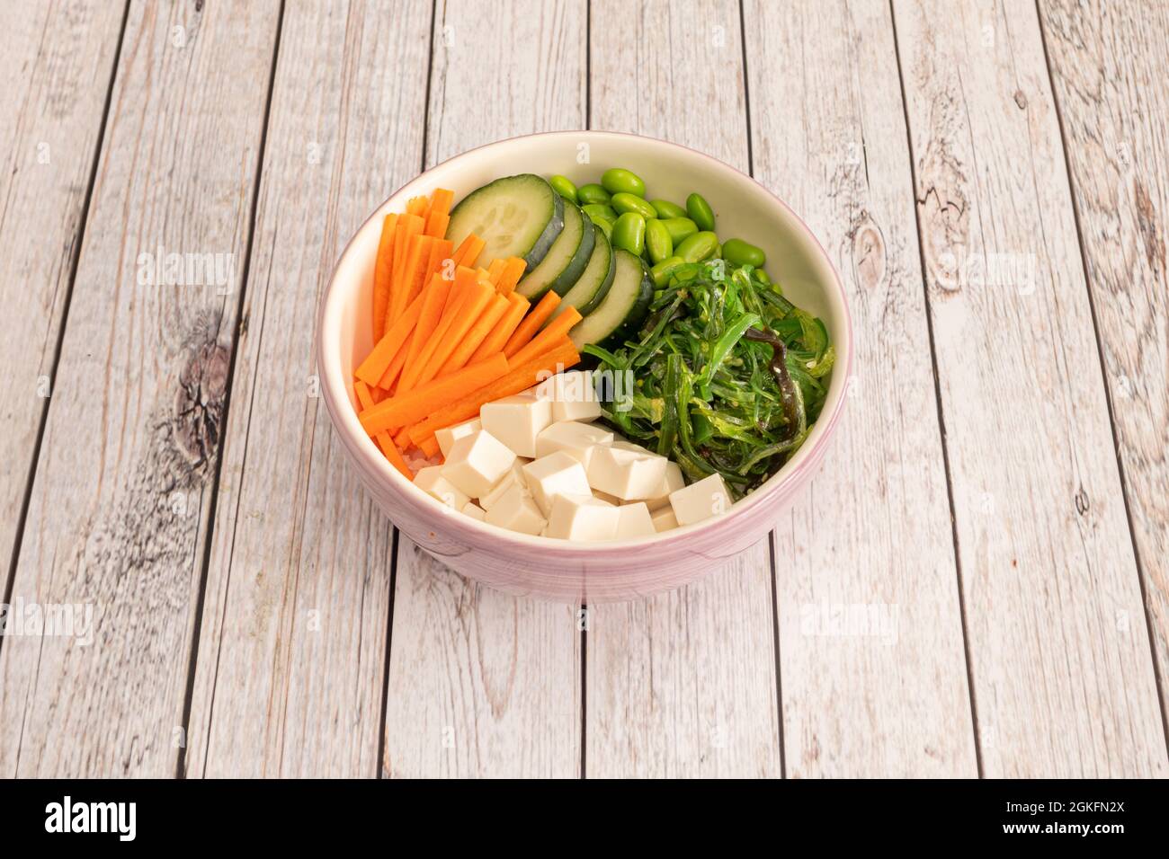 Ciotola di vegano rosa con insalata di alghe di wakame con tofu bianco a dadini, carota grattugiata, fette di cetriolo e fagioli di edamame su tavola di legno Foto Stock