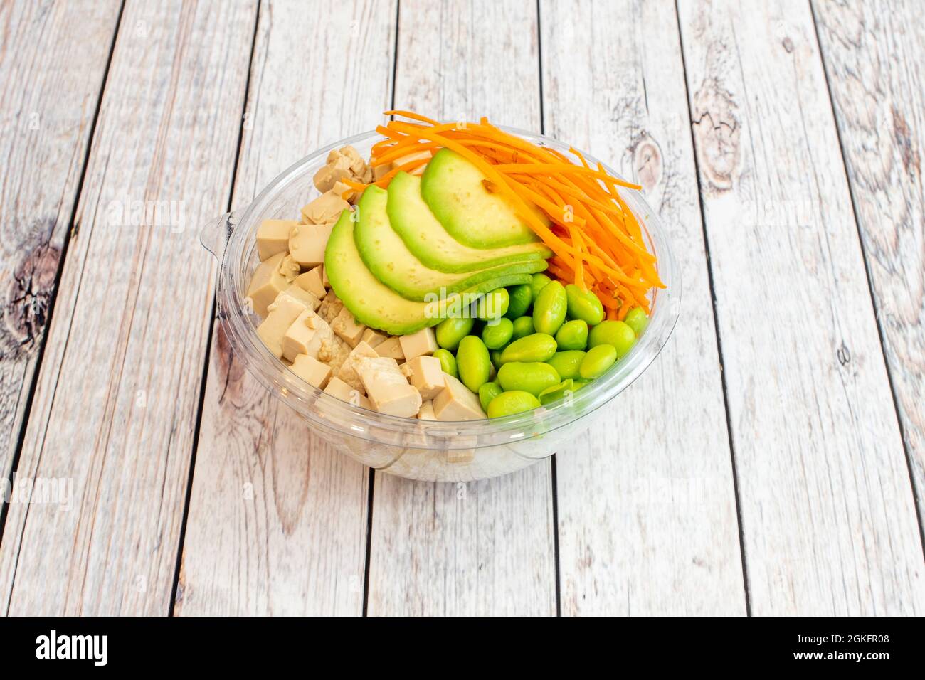 Ciotola sana di tofu senza gusto con fette di avocado maturo, scorza di carote, fagioli di edamame, base di riso bianco all'interno di una ciotola trasparente per la consegna a domicilio Foto Stock