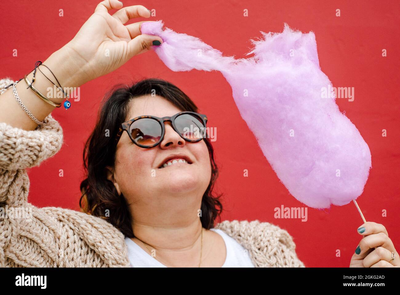 La donna strappa un pezzo di caramella di cotone lilla per mangiarlo, indossa bicchieri scuri ed è su sfondo rosso Foto Stock