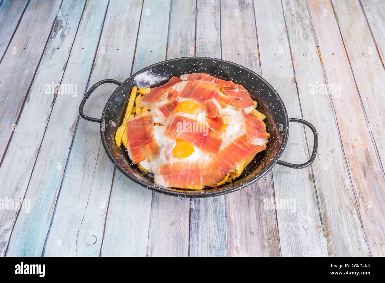 Padella in metallo smaltato nero con manici riempiti con una tipica ricetta  spagnola da condividere con uova rotte con prosciutto e patate fritte su  teglia di legno Foto stock - Alamy