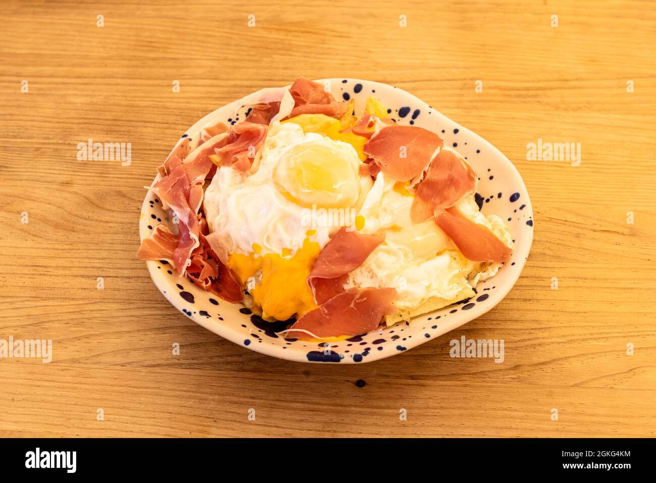 Famoso piatto spagnolo di uova rotte con prosciutto Serrano per condividere lo stile tapas su una ciotola bianca con puntini neri su un tavolo di legno Foto Stock