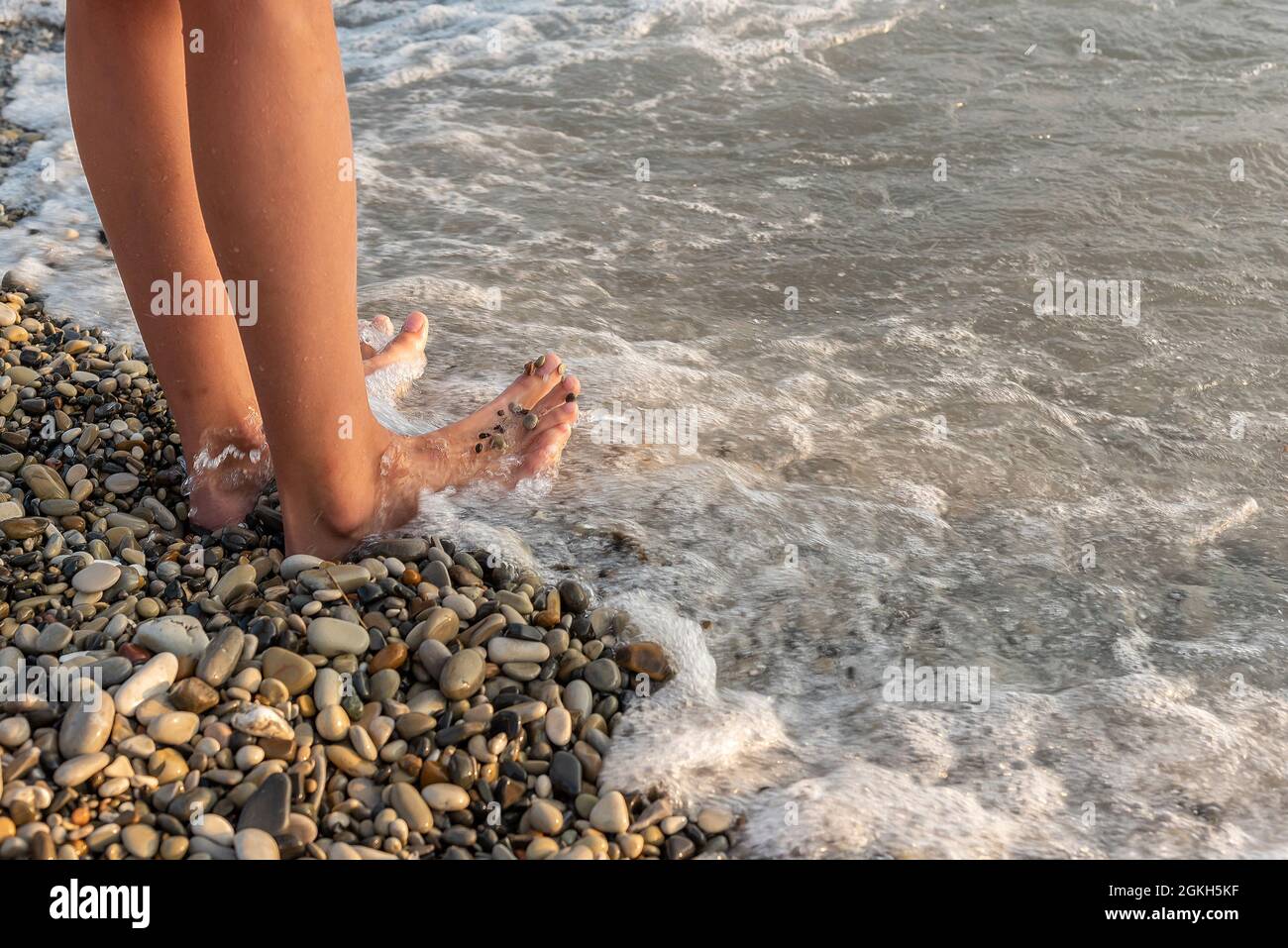 Donna a piedi nudi si erge in un'onda di mare schiumosa sulla spiaggia di ciottoli Foto Stock