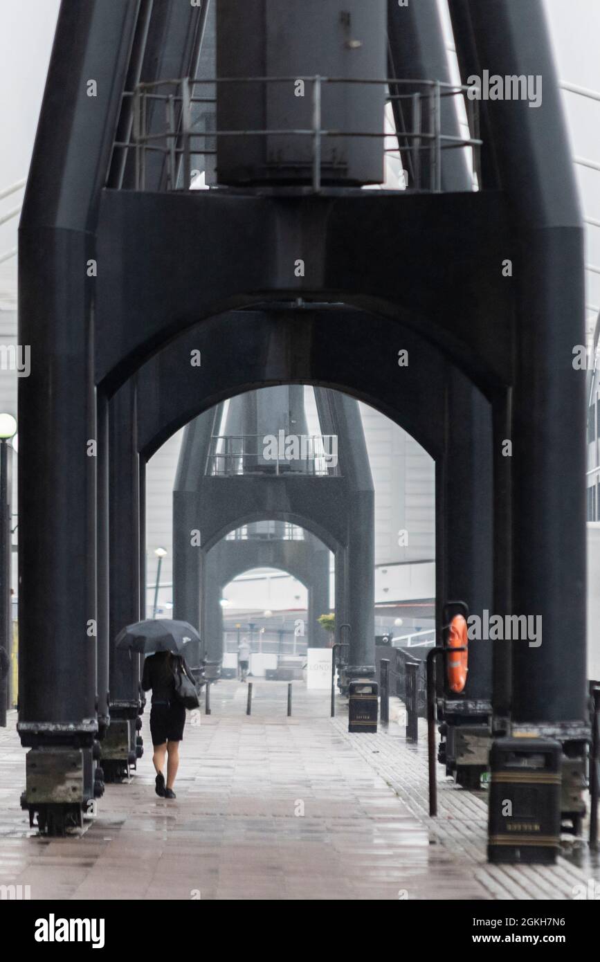 Donna che cammina sotto le enormi gru del molo presso il Royal Victoria Dock, Newham, Londra, Regno Unito, sotto la pioggia che tiene un ombrello. Brutto tempo estivo Foto Stock