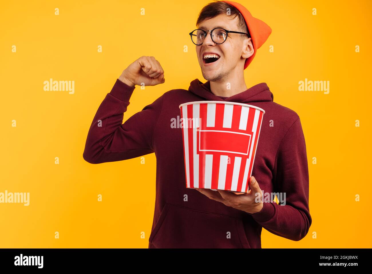 Ritratto di felice uomo eccitato che indossa occhiali e cappello rosso mostrando il gesto di vittoria e tenendo secchio di popcorn e mangiare popcorn su bac giallo Foto Stock