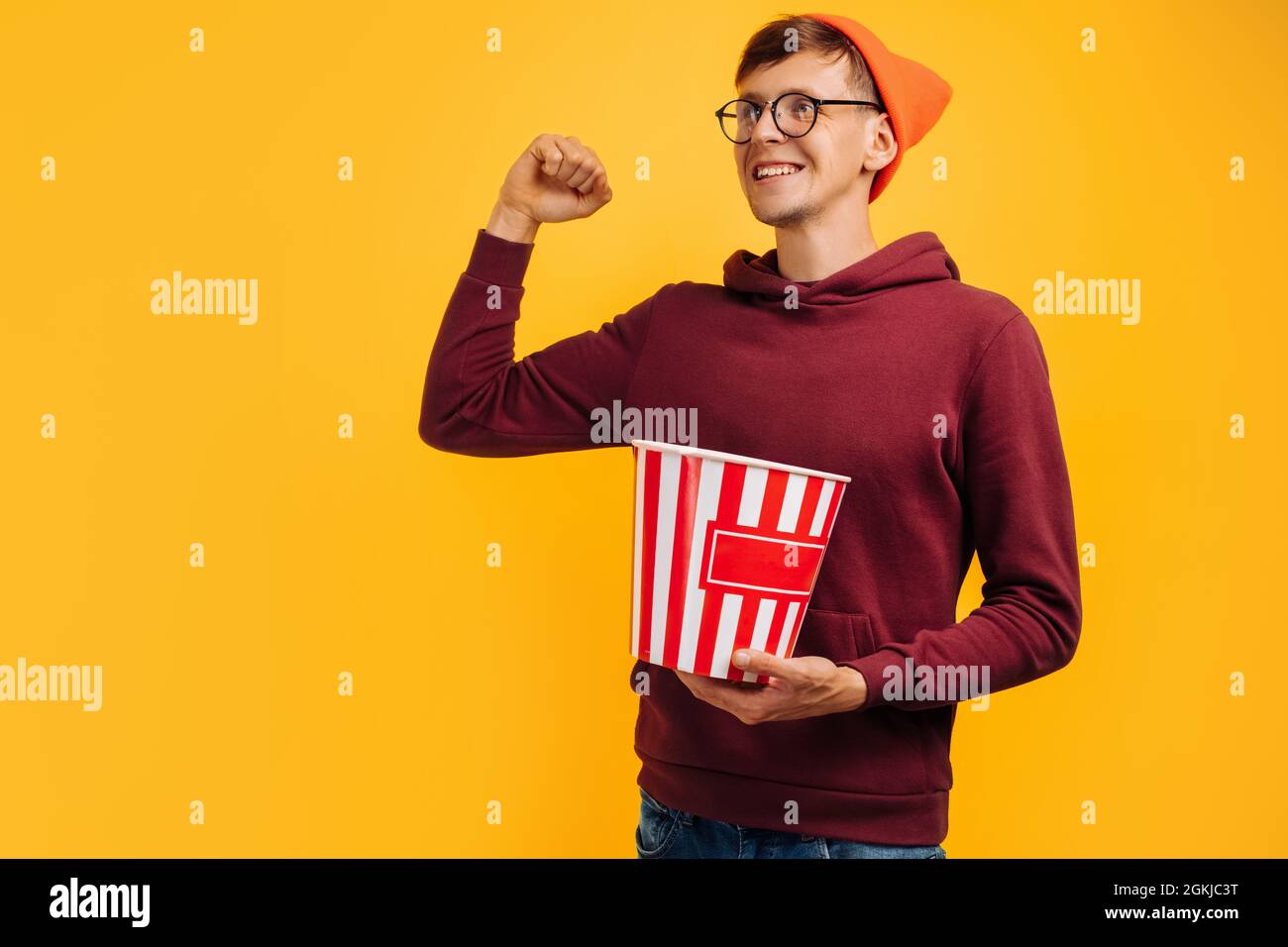 Ritratto di felice uomo eccitato che indossa occhiali e cappello rosso mostrando il gesto di vittoria e tenendo secchio di popcorn e mangiare popcorn su bac giallo Foto Stock