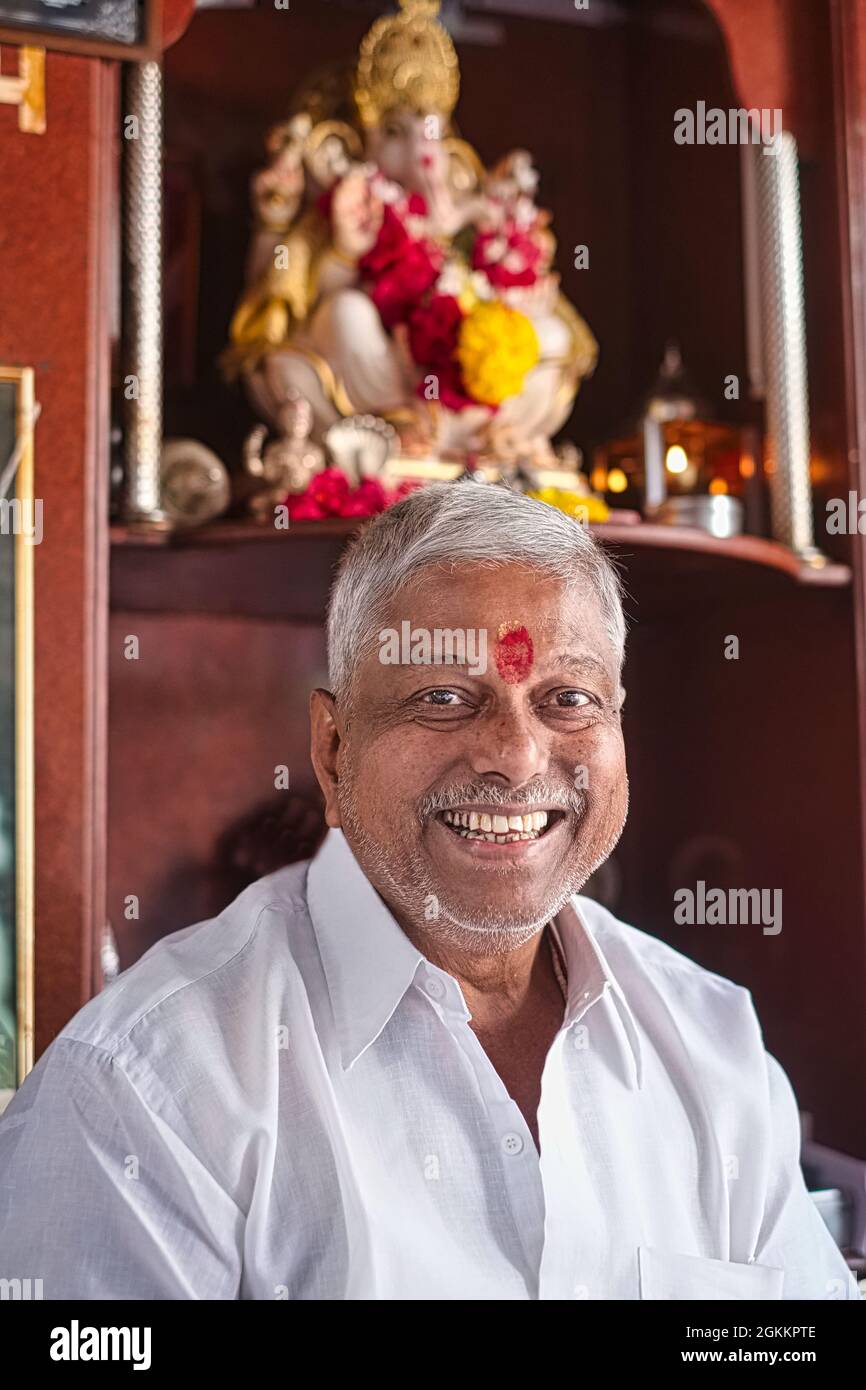 Un sorridente negoziante indù a Mumbai, in India, con un segno di tika rosso sulla fronte e una figura di Ganesh (dio elefante indù) dietro di lui Foto Stock
