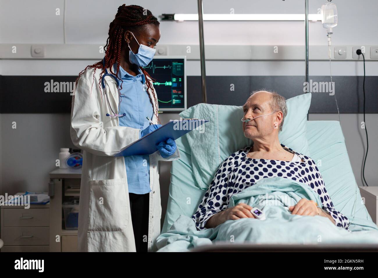 Medico afroamericano con maschera chirurgica che legge la diagnosi dalla clipboard a malato malato malato paziente anziano che si posa a letto, respirando con l'aiuto della maschera di ossigeno, ascoltando discutere con il personale medico circa il recupero. Foto Stock