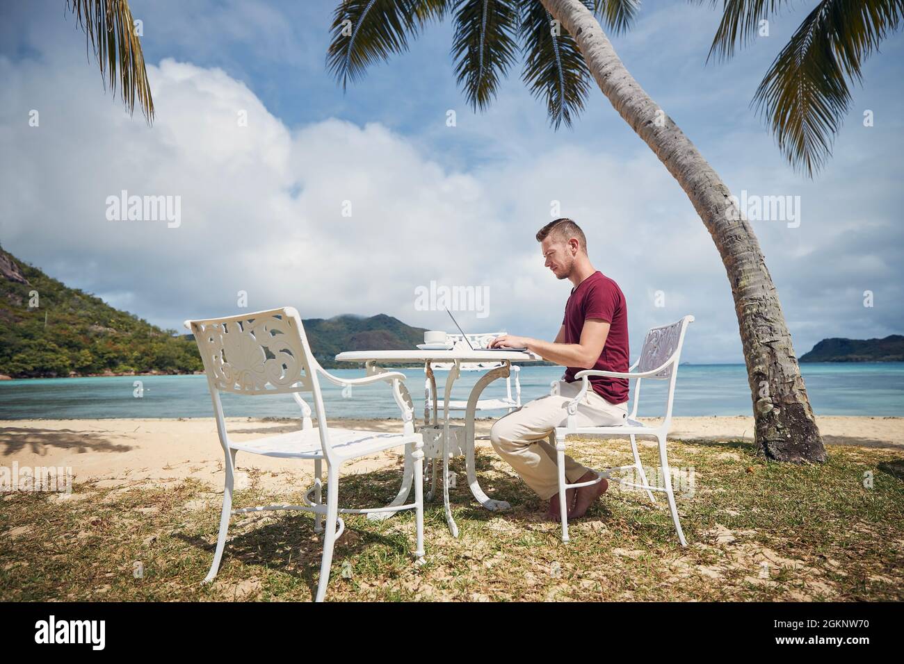 Giovane freelance che lavora su un notebook da una destinazione tropicale. Uomo seduto sotto le palme sulla spiaggia. Foto Stock
