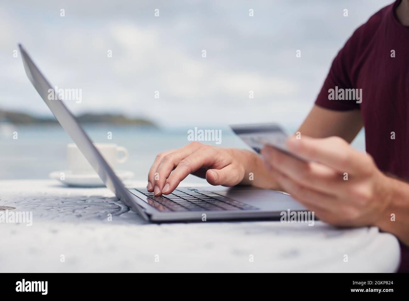 Uomo seduto sulla spiaggia contro il mare e utilizzando la carta di credito per la prenotazione online o lo shopping via computer portatile. Foto Stock