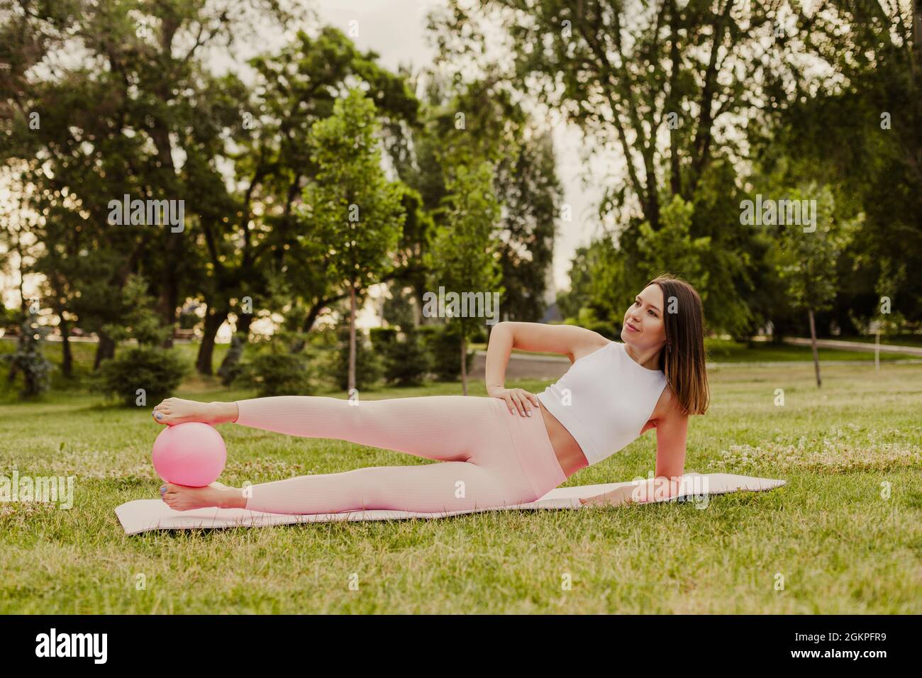 giovane, bella donna in rosa sportswear è sdraiata su tappeto sportivo di lato con pilates palla bloccato tra i piedi sul prato nel parco all'alba Foto Stock
