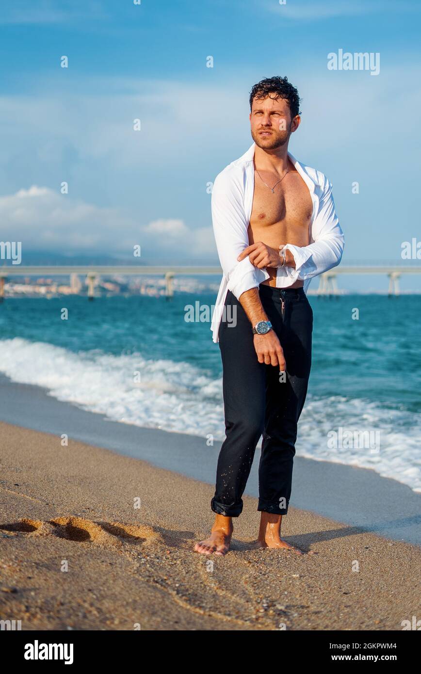 Uomo elegante e spensierato con corpo muscolare in piedi sulla spiaggia vicino al mare in serata e guardando via Foto Stock