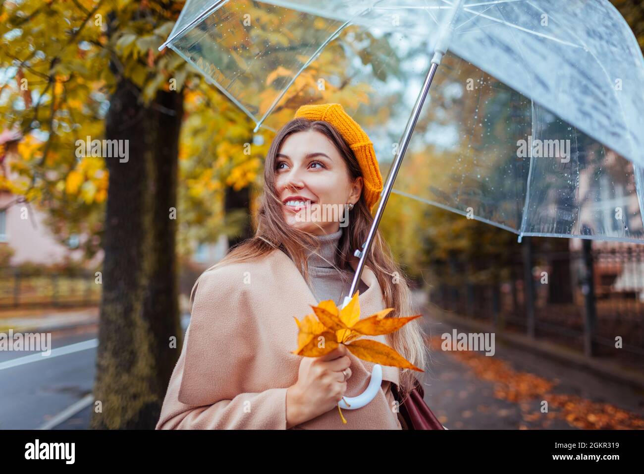 Felice giovane donna che cammina all'aperto sotto ombrello trasparente  durante la pioggia tenendo foglie gialle. Ragazza elegante che indossa  abiti e accessori alla moda Foto stock - Alamy