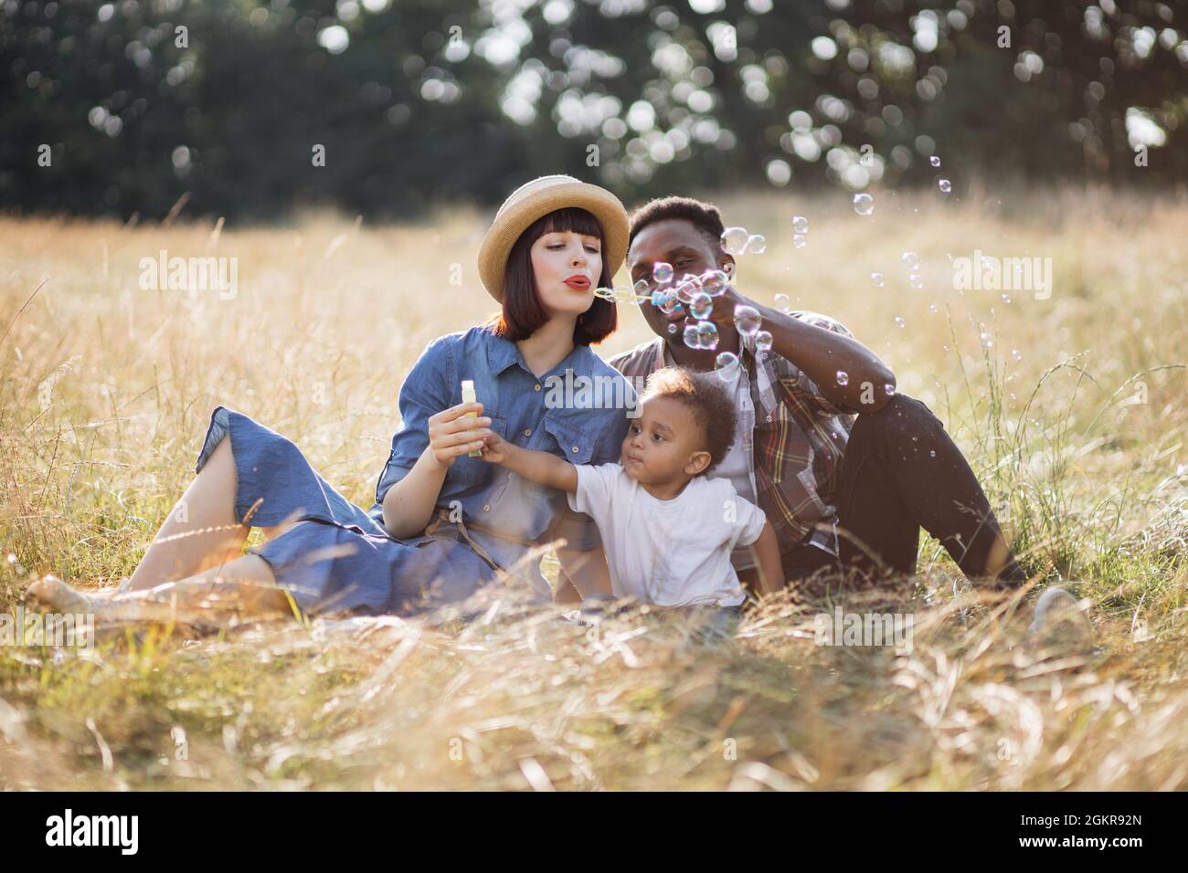 Giovani genitori multiculturali che usano le bolle di sapone mentre giocano con il figlio piccolo all'aperto. Famiglia felice seduto sul prato e godere di tempo libero durante i giorni estivi. Foto Stock