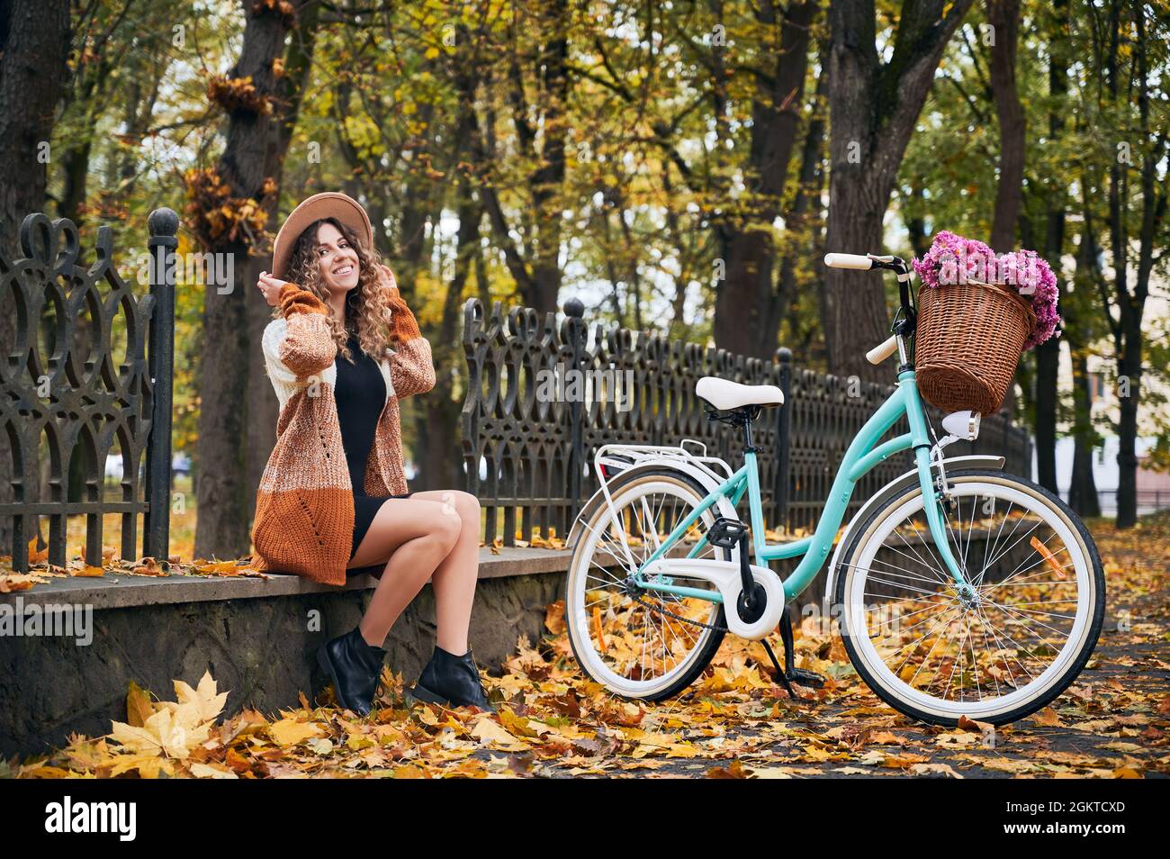 Donna positiva seduta su calcestruzzo bordesure di recinto parco vicino alla sua bici di città blu brillante femmina a caldo giorno d'autunno. Attività ricreative attive, giro in bicicletta negli spazi aperti della città. Foto Stock