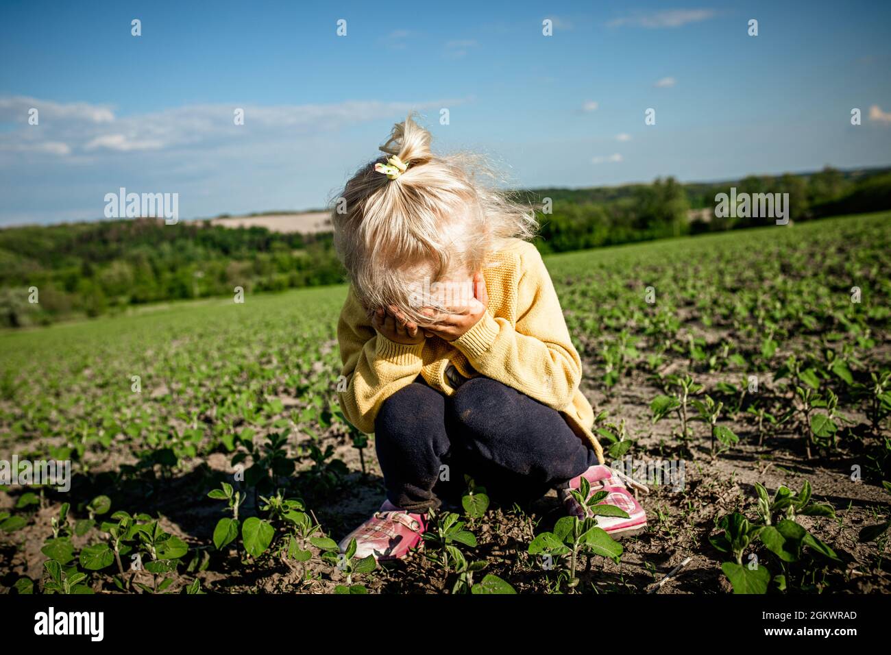 il bambino triste si siede sul campo, squattando e grida con gli occhi ricoperti di mani. Piccola bionda seria ragazza stanca che squatting da solo in un campo vuoto di giovani Foto Stock